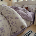 A cozy bed made with a floral duvet cover and matching pillowcases in lavender and cream, with a solid lavender sheet underneath. A small teddy bear sits on the pillow in a bright bedroom setting.

