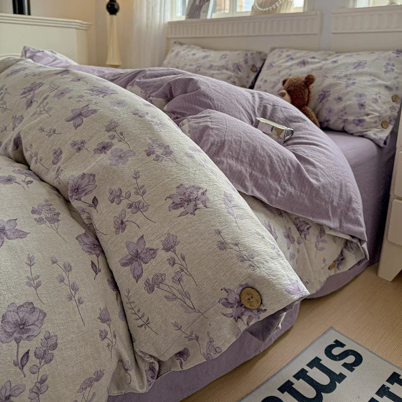 A cozy bed made with a floral duvet cover and matching pillowcases in lavender and cream, with a solid lavender sheet underneath. A small teddy bear sits on the pillow in a bright bedroom setting.

