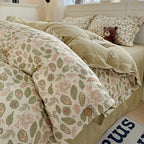 A cozy bed dressed in a cream-colored duvet cover and matching pillowcases featuring a botanical leaf pattern in sage green and dusty pink, with solid sage green pillow shams and a small brown teddy bear resting on the bed.

