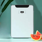 A white rectangular countertop appliance with rounded corners, a black top panel, and a small digital temperature display reading 26°C. The appliance is photographed on a light surface against a soft teal background, with green leaves at the upper left and watermelon slices at the lower right.

