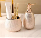 A bathroom or vanity set featuring a textured metallic gold toothbrush holder with a white toothpaste tube and two gold toothbrushes, alongside a matching textured gold pump bottle with a rose gold-toned dispenser cap, set against a warm beige background.

