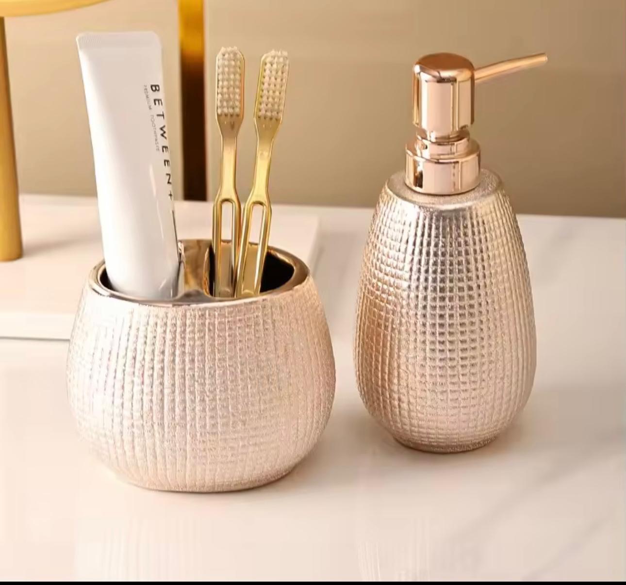 A bathroom or vanity set featuring a textured metallic gold toothbrush holder with a white toothpaste tube and two gold toothbrushes, alongside a matching textured gold pump bottle with a rose gold-toned dispenser cap, set against a warm beige background.

