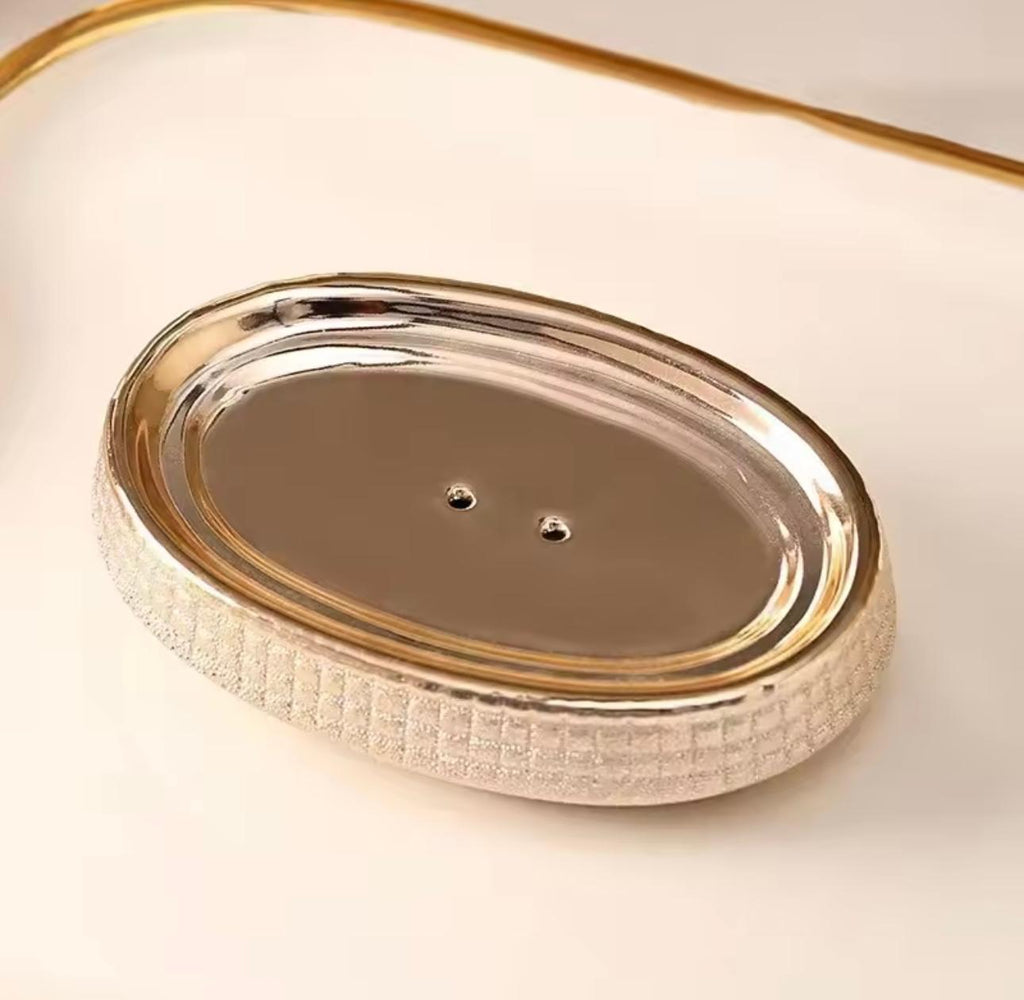 An oval metallic soap dish with a textured, woven-look silver border and a polished gold-toned rim, shown on a warm cream background.

