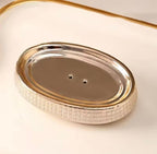 An oval metallic soap dish with a textured, woven-look silver border and a polished gold-toned rim, shown on a warm cream background.

