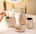 A bathroom toiletry set in a warm metallic champagne finish, including a textured tooth holder with toothbrushes, a pump bottle, a cup, and a lid, arranged on a white vanity next to a gold-framed mirror.


