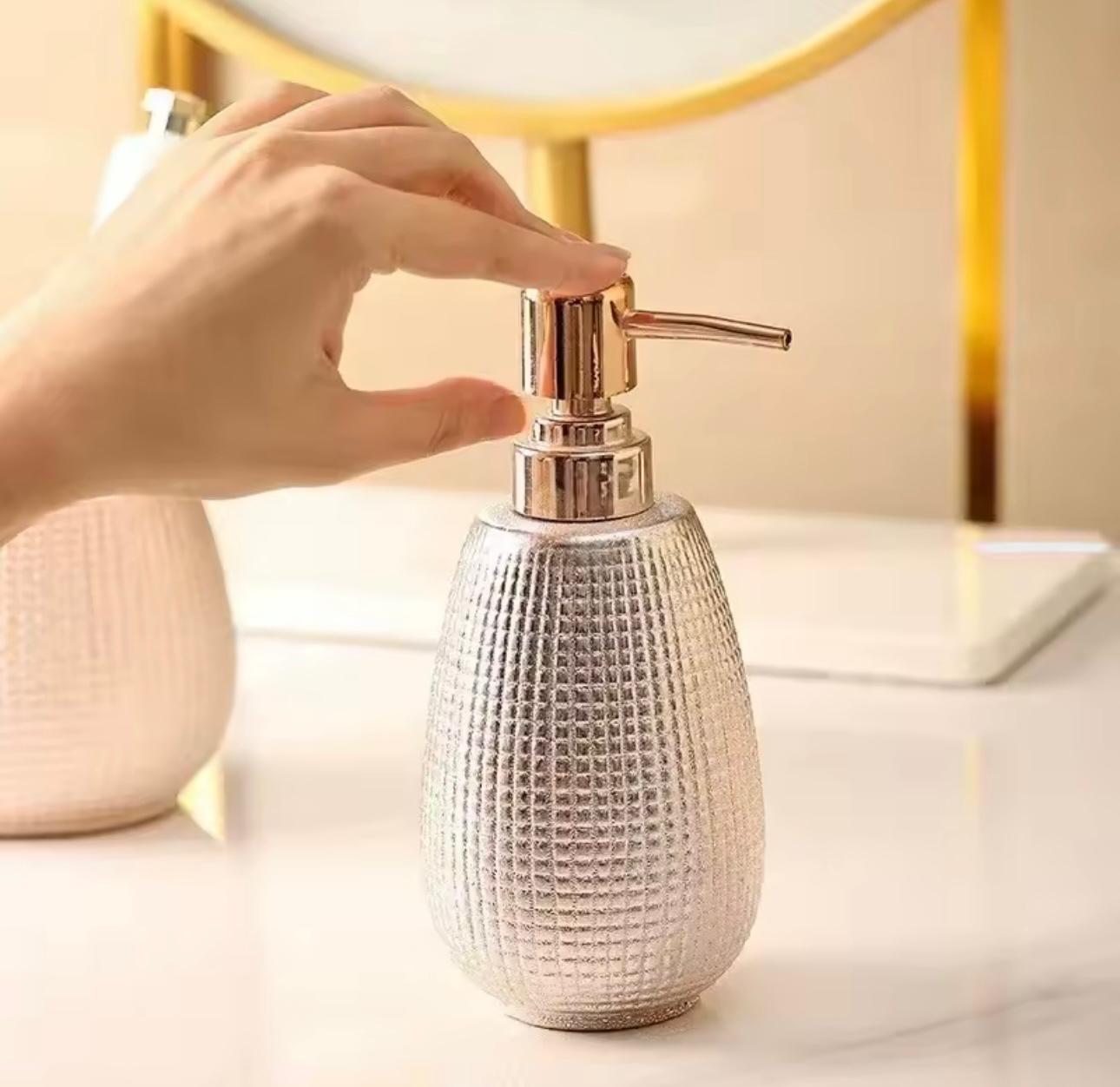 A hand is shown pressing the pump dispenser of a textured metallic silver soap or lotion bottle with a rose gold pump, set on a light marble countertop.

