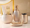 A coordinated bathroom or toothbrush holder set in a textured metallic finish, including a round cup with toothbrushes and a tube, a pump bottle, a small cup, and a round lid, arranged on a light surface against a soft beige background.

