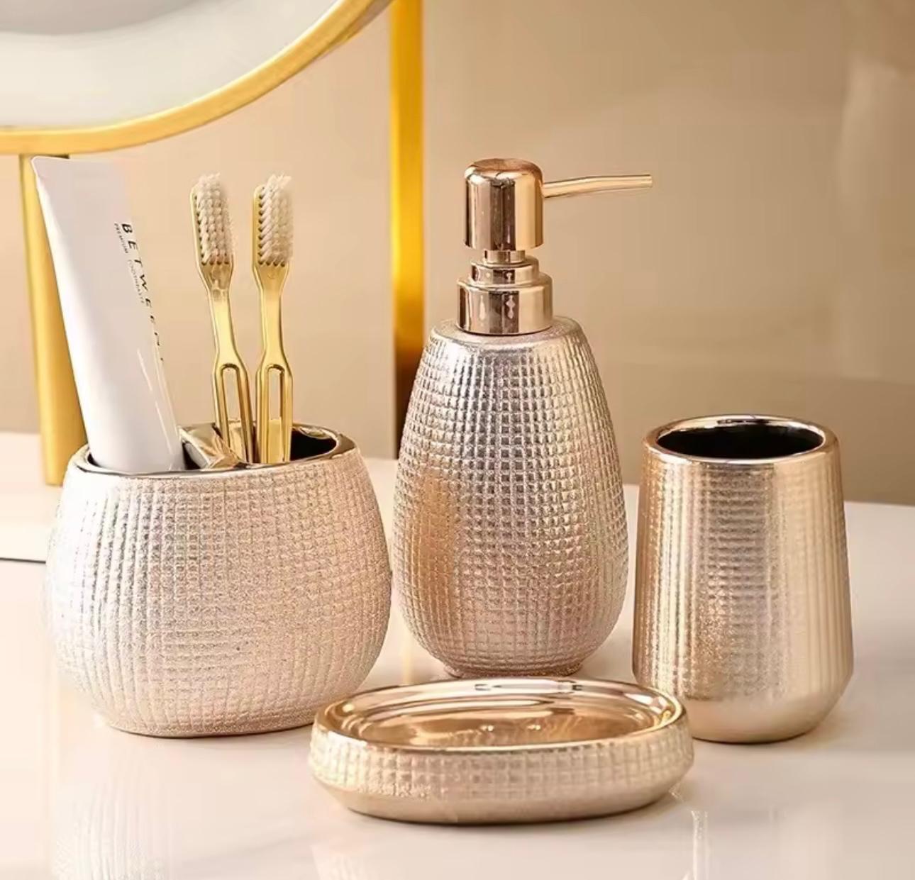 A coordinated bathroom or toothbrush holder set in a textured metallic finish, including a round cup with toothbrushes and a tube, a pump bottle, a small cup, and a round lid, arranged on a light surface against a soft beige background.


