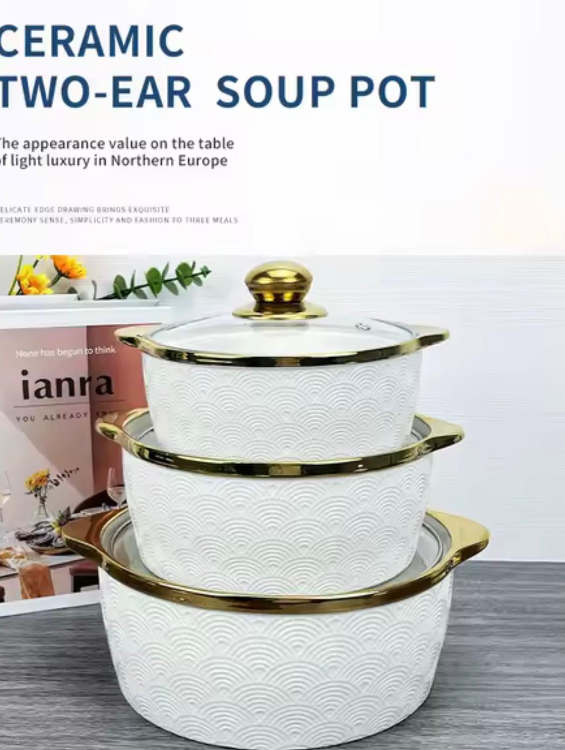 Three stacked ceramic two-ear soup pots with lids in white with gold trim and handles, set on a gray surface with a magazine in the background.

