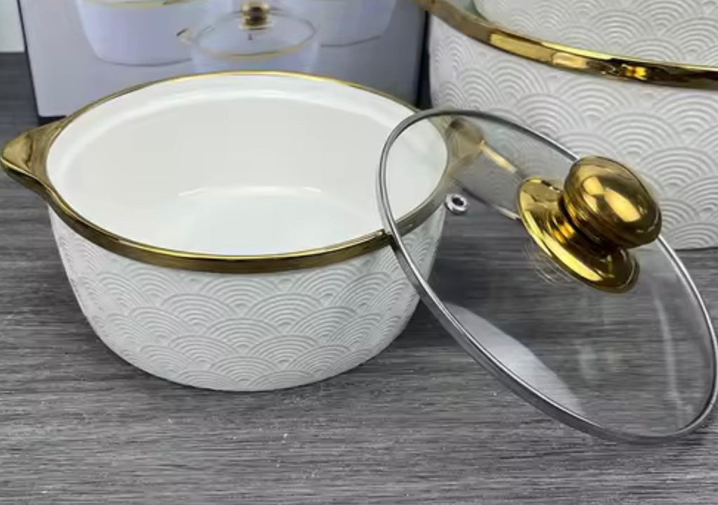 A white ceramic stockpot with a gold rim and a matching glass lid featuring a gold knob, displayed on a gray wood-textured surface.

