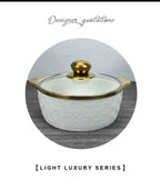 A white ceramic lidded casserole dish with a gold-trimmed glass lid and gold knob, shown inside a circular frame on a gray wood-textured background with decorative text above and below.

