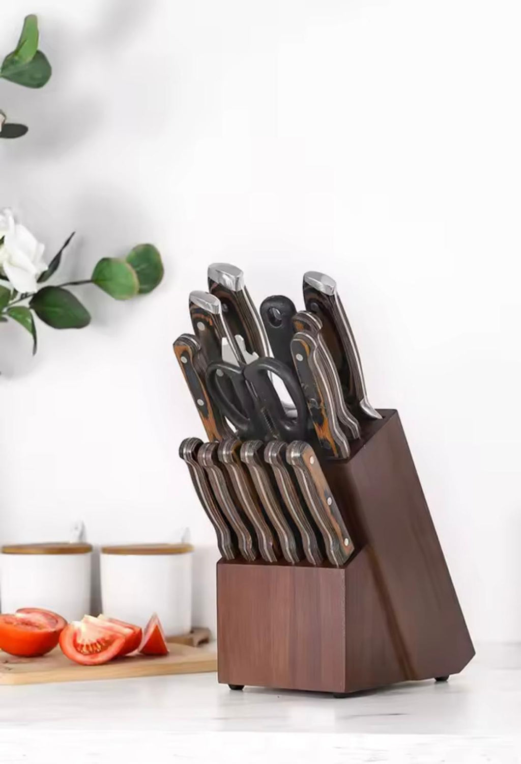 A wooden knife block filled with kitchen knives and scissors, set on a countertop with a blurred kitchen background and tomato slices nearby.

