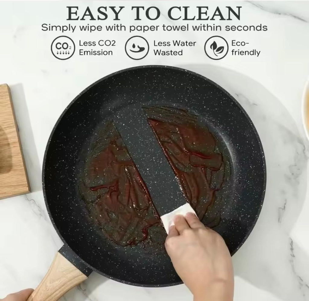 A black non-stick frying pan with a wooden handle sits on a white marble surface, showing dark residue inside. A hand wipes the pan with a paper towel, and marketing text at the top reads 'EASY TO CLEAN' with icons for less CO2 emission, less water wasted, and eco-friendly.

