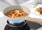 A white non-stick frying pan with a light wood handle sits on a black gas stove burner, cooking a seafood pasta dish with shrimp, lemon, and tomatoes. A plate of food is visible in the background on a white countertop.

