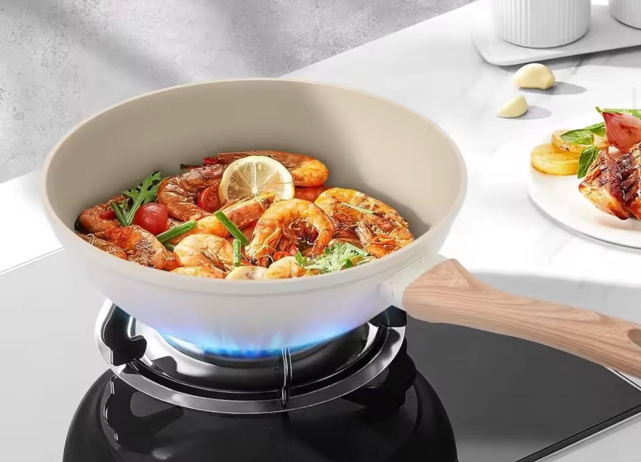 A white non-stick frying pan with a light wood handle sits on a black gas stove burner, cooking a seafood pasta dish with shrimp, lemon, and tomatoes. A plate of food is visible in the background on a white countertop.

