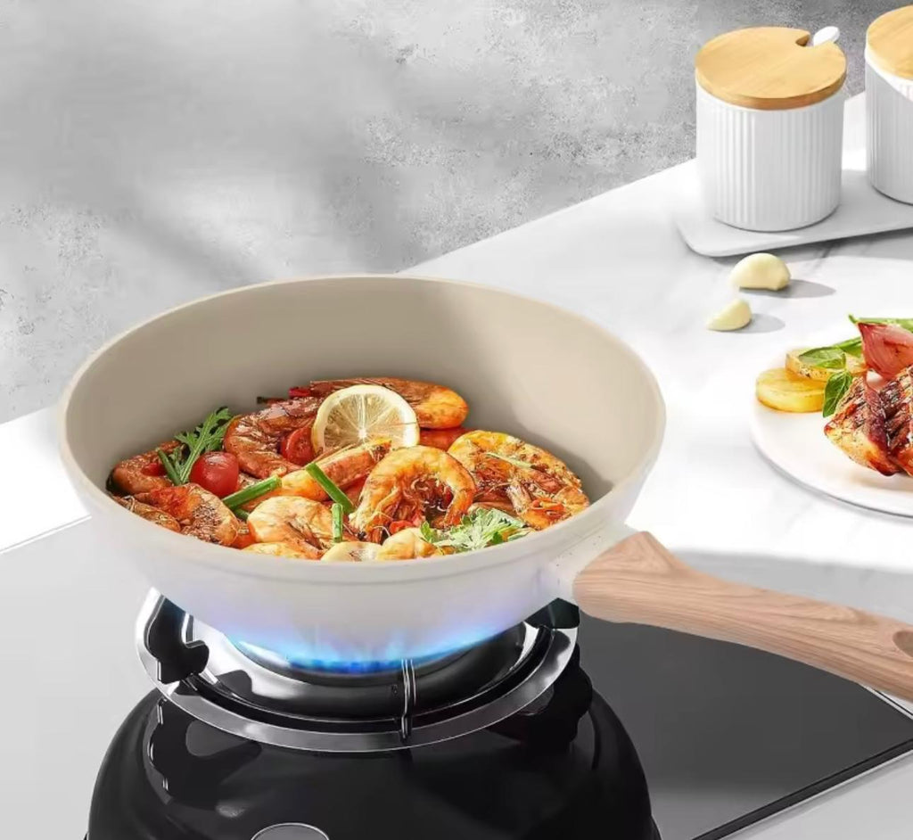 A beige ceramic non-stick pan with a wooden handle sits on a black gas stove burner, cooking a seafood dish with shrimp, tomatoes, and a lemon slice. The background is a clean, modern kitchen setting with white marble countertops and white ceramic canisters.

