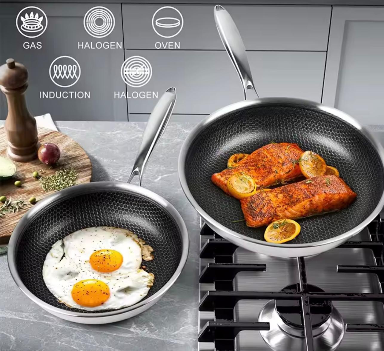 Two non-stick frying pans with silver handles and bases, shown on a stovetop and counter in a kitchen setting. One pan contains two sunny eggs, and the other contains salmon fillets with lemon slices.

