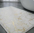 A rectangular bath mat with a white marble pattern and gold veins, shown on a gray floor next to a white bathtub.

