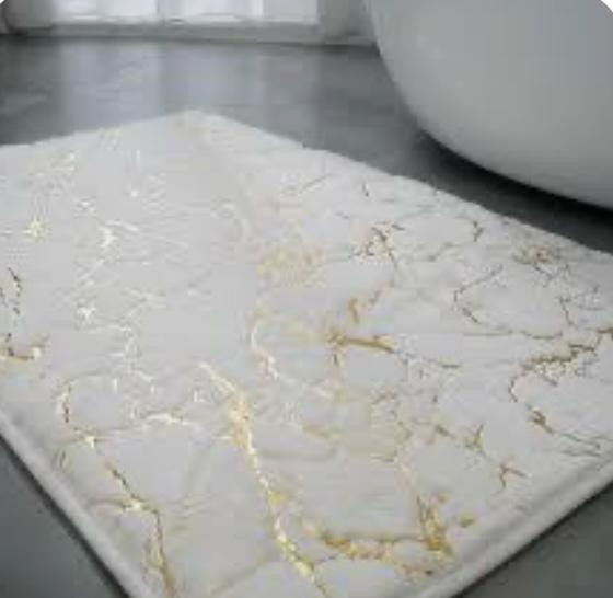 A rectangular bath mat with a white marble pattern and gold veins, shown on a gray floor next to a white bathtub.

