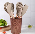A silicone kitchen utensil set in a cylindrical wooden holder, photographed on a kitchen counter with vegetables and a small bowl in the background. The utensils are light beige with rose gold handles.

