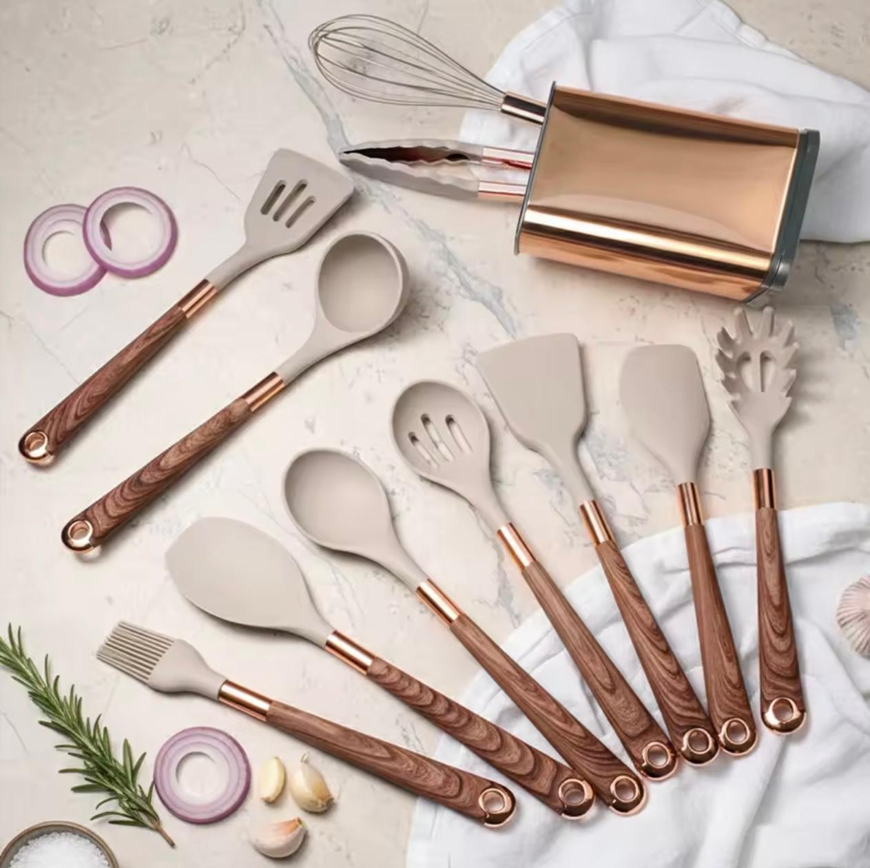 A 12-piece silicone kitchen utensil set with rose gold metal accents and wood-grain handles, arranged on a light marble surface with food props like onion, garlic, rosemary, and a whisk.

