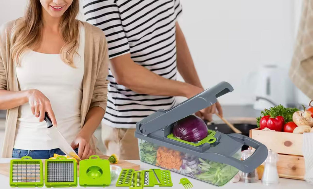 A kitchen scene showing a manual vegetable slicer and dicer set in use by a couple. The gray and bright green plastic slicer is loaded with an onion and other vegetables, while a woman in a white top and beige cardigan holds a knife, and a man in a black-and-white striped shirt stands behind her.

