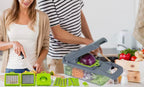 A kitchen scene showing a manual vegetable slicer and dicer set in use by a couple. The gray and bright green plastic slicer is loaded with an onion and other vegetables, while a woman in a white top and beige cardigan holds a knife, and a man in a black-and-white striped shirt stands behind her.

