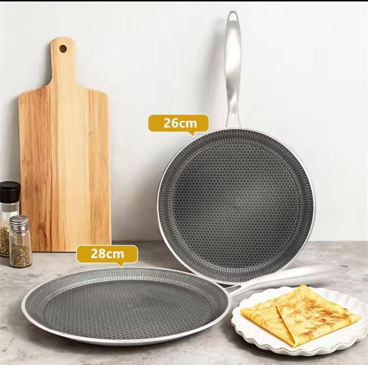 A set of non-stick cookware shown on a countertop against a light wall: a rectangular wooden cutting board, a frying pan labeled 26cm, a shallow round pan labeled 28cm, and a small plate with folded pancakes. Yellow size callouts indicate the pan diameters.

