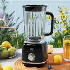 A black countertop blender with a clear pitcher and black handle, shown on a wooden tray outdoors with lemons, a glass of orange drink, and blurred flowers in the background.

