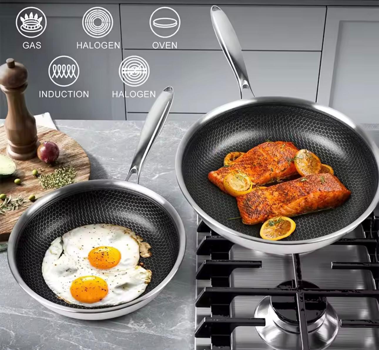Two nonstick frying pans with textured interiors are shown on a stovetop in a kitchen setting. One pan contains fried eggs, and the other contains salmon fillets with lemon slices.

