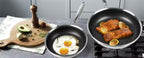 Two black non-stick frying pans on a stovetop in a kitchen setting, one frying eggs and the other cooking salmon with lemon slices, alongside a cutting board with avocado and spices.

