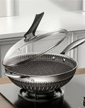 A stainless steel wok with a black handle and a clear glass lid, shown on a stovetop in a kitchen setting.

