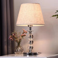 Table lamp with a textured shade and glass base next to a vase with flowers on a surface.

