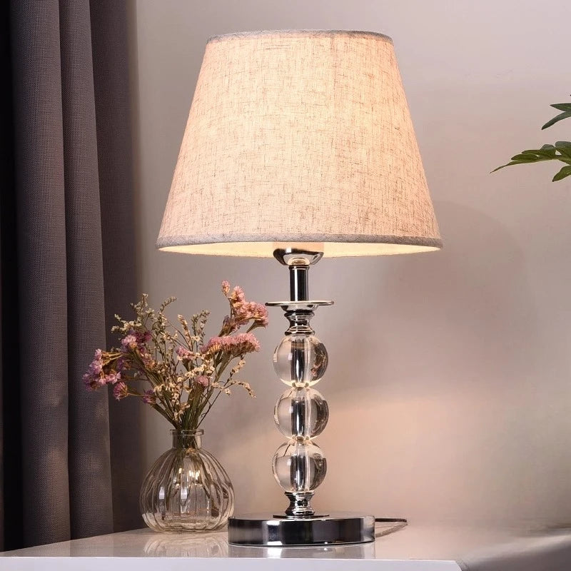 Table lamp with a textured shade and glass base next to a vase with flowers on a surface.

