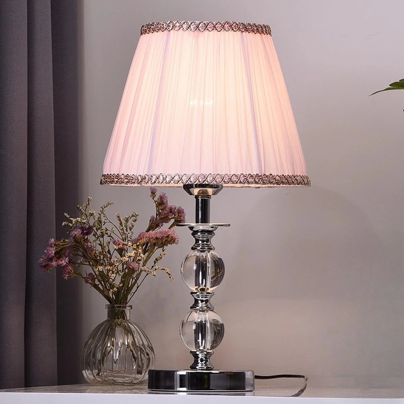 Decorative table lamp with a pleated white shade and glass base on a surface.

