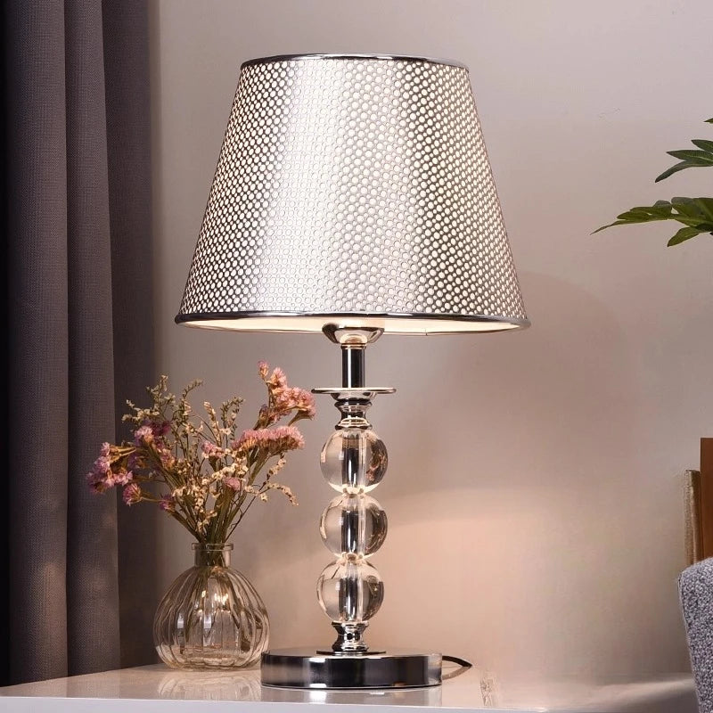 A table lamp with a textured silver and white patterned drum shade, a clear glass beaded column base, and a polished silver metal stem, shown on a tabletop next to a glass vase with dried pink flowers.

